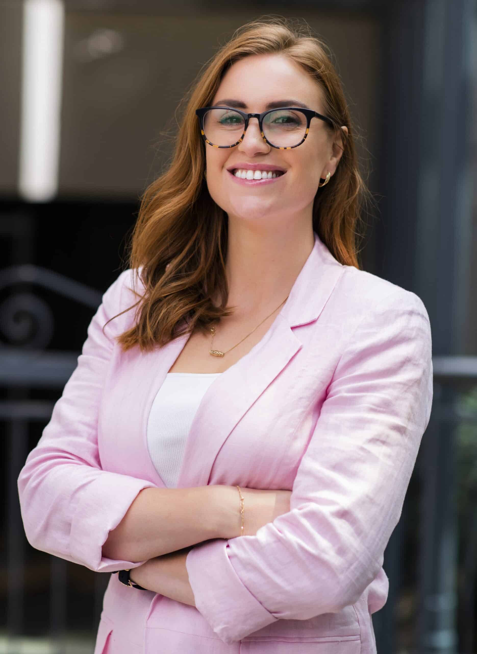 A woman, elegantly dressed in a pink suit and glasses, captured in a professional headshot photograph